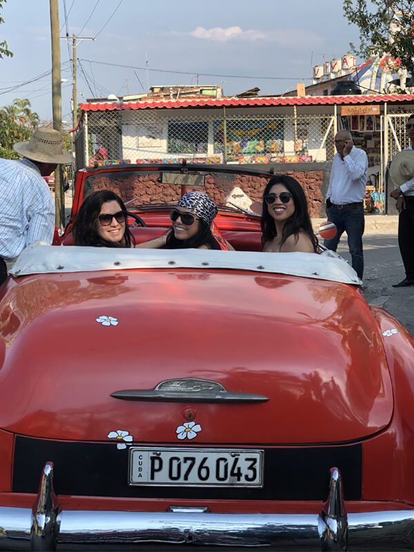 Three women in a red car with people standing around.